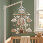 A crocheted baby mobile with a hot air balloon, clouds, stars, and a teddy bear in a basket, hanging from a white crib bar in a nursery with a sage green wall and wooden crib.