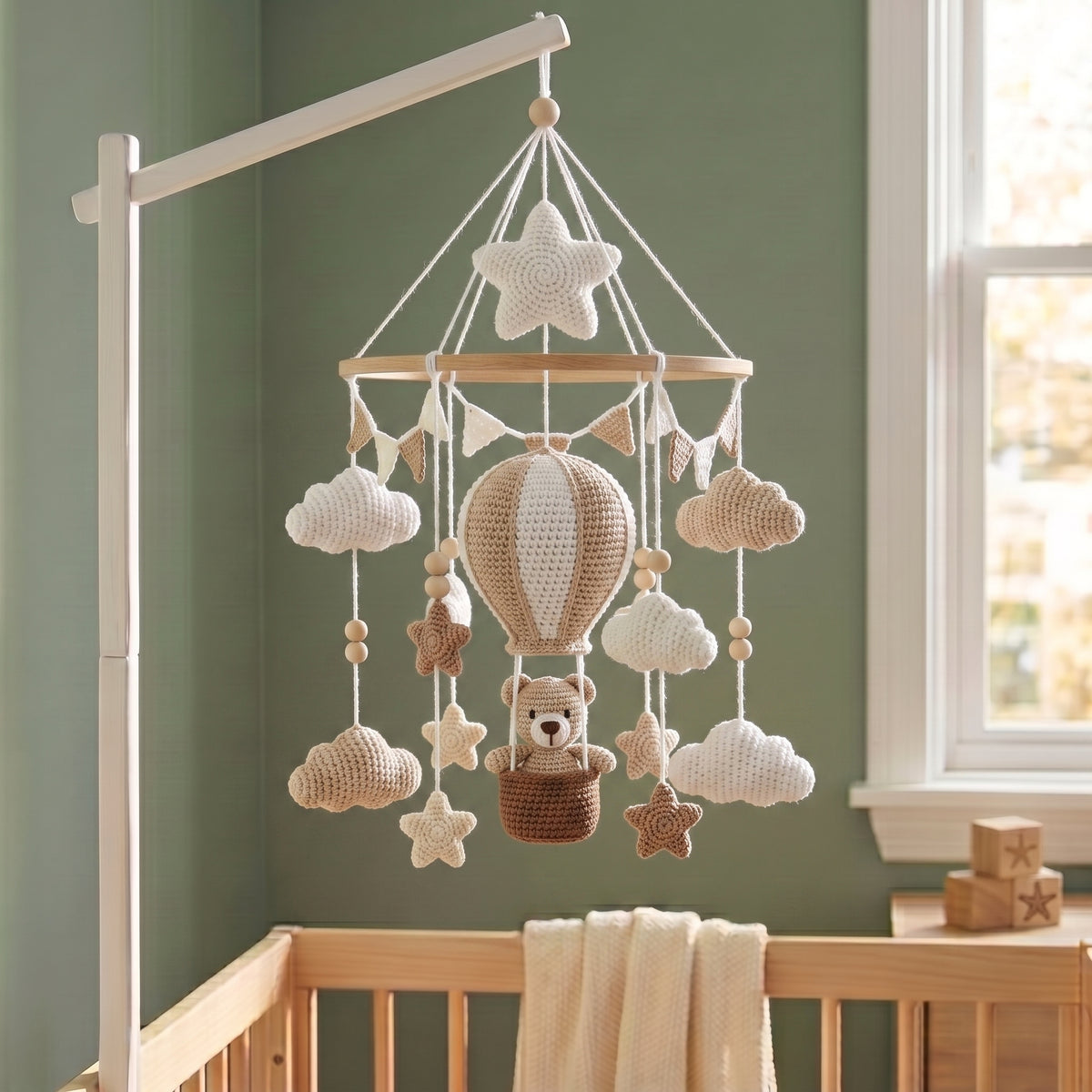 A crocheted baby mobile with a hot air balloon, clouds, stars, and a teddy bear in a basket, hanging from a white crib bar in a nursery with a sage green wall and wooden crib.