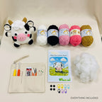 A crocheted plush cow kit laid out on a cream fabric background, including a finished cow plush toy, five balls of yarn in white, black, pink, magenta, and mustard, a printed instruction booklet, a canvas tool pouch with crochet hooks and accessories, loose white stuffing, and small stitch markers. Text at the bottom reads 'EVERYTHING INCLUDED'.