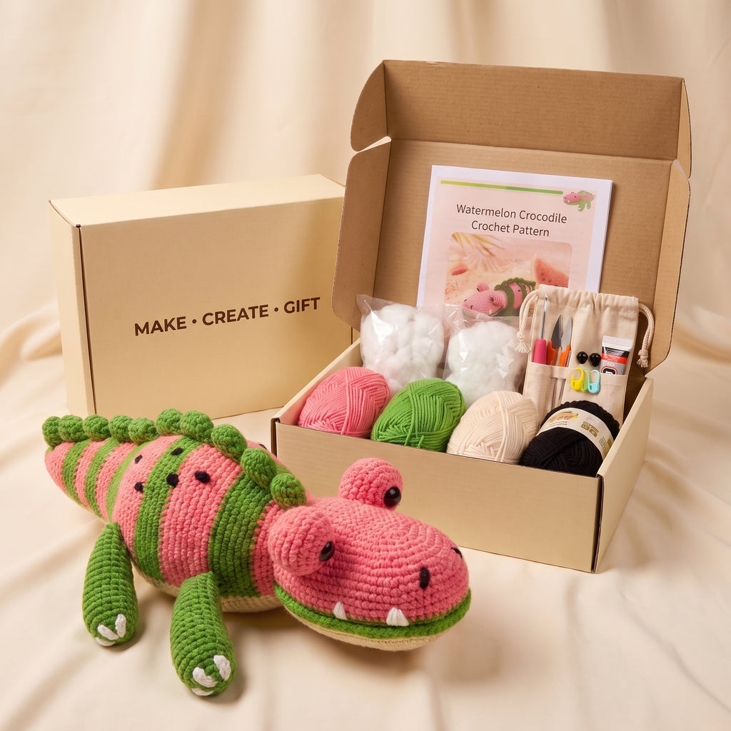 A crocheted watermelon crocodile toy is displayed in front of an open kraft gift box containing a crochet pattern card, yarn balls in pink, green, cream, and black, and a small crochet needle pouch. A closed kraft box with the text 'MAKE • CREATE • GIFT' is visible behind it on a beige fabric backdrop.