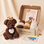 A crocheted puppy toy sits beside an open cardboard gift box containing a crochet pattern booklet, two balls of yarn, a drawstring bag, and small crafting tools on a cream fabric backdrop.