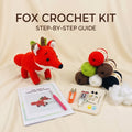 A fox crochet kit laid out on a beige fabric background with the text 'FOX CROCHET KIT' and 'STEP-BY-STEP GUIDE' at the top. The kit includes a crocheted red fox toy, a crochet pattern booklet, a pink crochet hook, thread, buttons, clips, glue, stuffing, and multiple yarn balls in red, green, brown, white, and black.