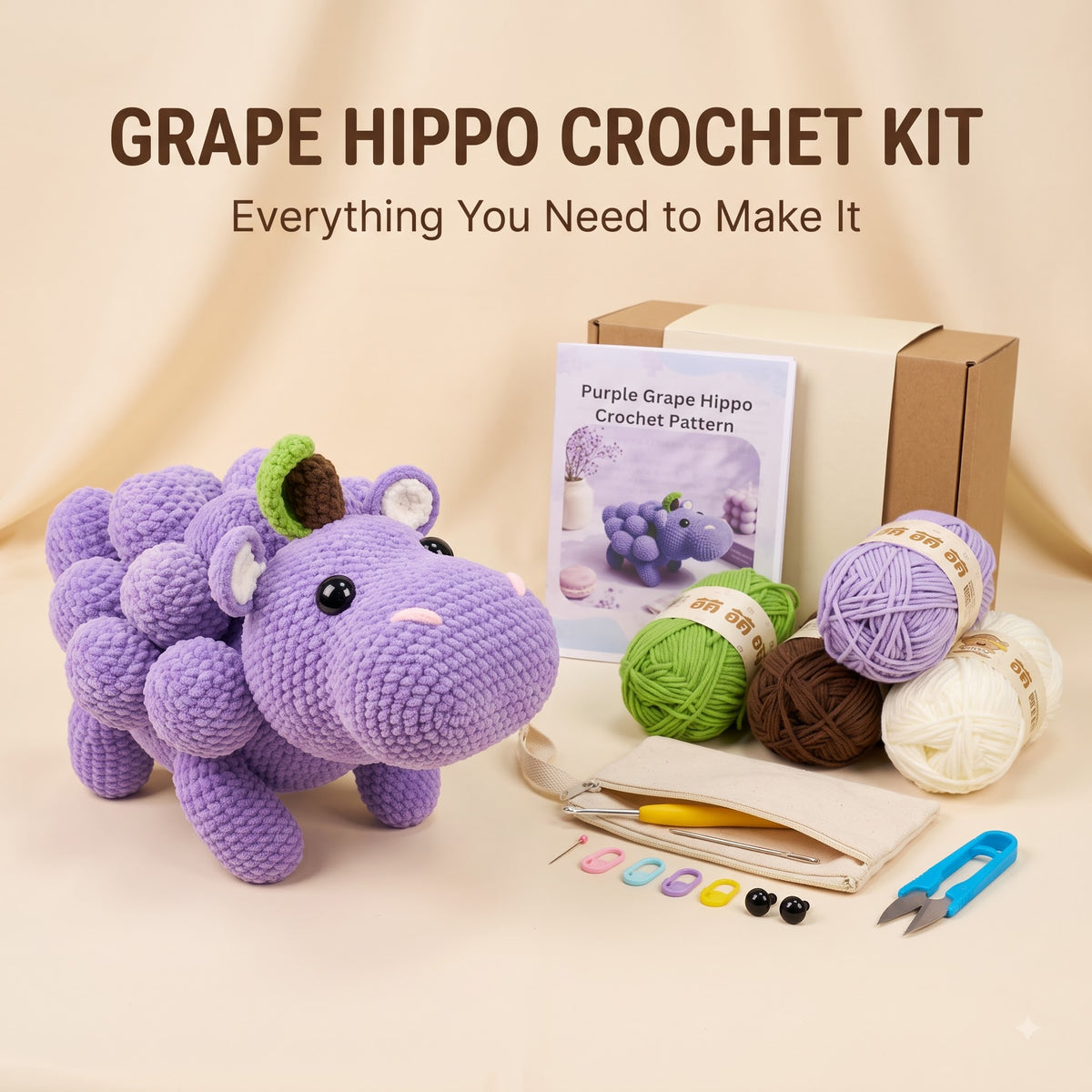 Grape Hippo Crochet Kit displayed on a beige fabric background, featuring a crocheted purple grape hippo toy, a crochet pattern booklet, a cardboard box, yarn balls in purple, green, brown, and cream, a crochet hook, buttons, and scissors.
