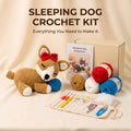 Sleeping Dog Crochet Kit displayed on a beige fabric backdrop, featuring a crocheted sleeping dog toy with a red bow and blue ball, a booklet, a kraft box, yarn balls in red, blue, white, and tan, crochet hooks in a fabric roll, and loose white fiber.