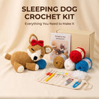 Sleeping Dog Crochet Kit displayed on a beige fabric backdrop, featuring a crocheted sleeping dog toy with a red bow and blue ball, a booklet, a kraft box, yarn balls in red, blue, white, and tan, crochet hooks in a fabric roll, and loose white fiber.