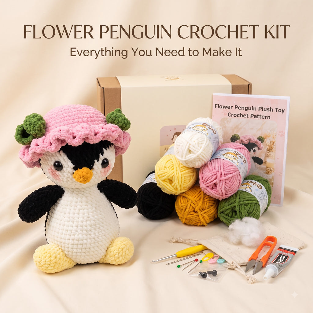 A crocheted penguin plush toy wearing a pink flower hat is displayed with a cardboard box, a crochet pattern booklet, multiple balls of yarn in black, yellow, pink, and green, and crochet tools on a beige fabric background. Text at the top reads 'FLOWER PENGUIN CROCHET KIT' and 'Everything You Need to Make It'.