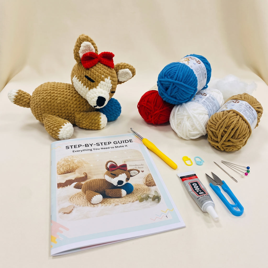 A crocheted fox toy kit displayed on a cream fabric background, featuring a finished tan and white crochet fox with a red bow, a printed step-by-step guide, a yellow crochet hook, blue and red yarn balls, white yarn, tan yarn, pins, glue, and other crafting tools.