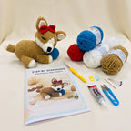 A crocheted fox toy kit displayed on a cream fabric background, featuring a finished tan and white crochet fox with a red bow, a printed step-by-step guide, a yellow crochet hook, blue and red yarn balls, white yarn, tan yarn, pins, glue, and other crafting tools.