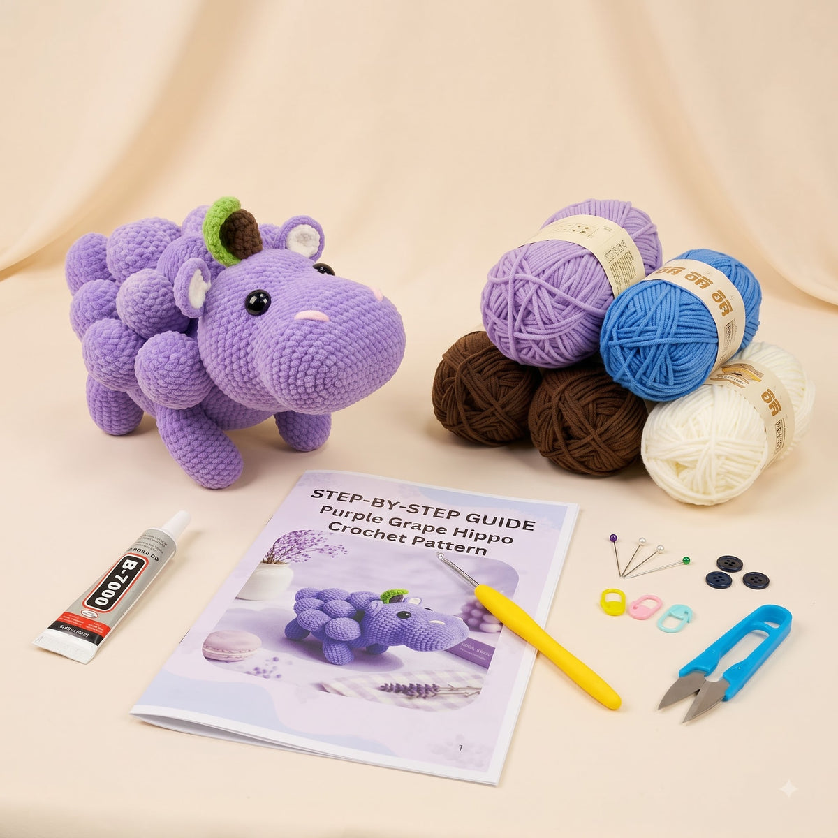 Crochet pattern kit for a purple grape hippo amigurumi doll, featuring a finished purple crocheted hippo, multiple balls of yarn in purple, blue, brown, and cream, a step-by-step guide booklet, a yellow crochet hook, buttons, pins, and a glue tube on a beige fabric background.