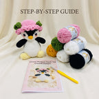 Crochet pattern kit for a Flower Penguin Plush Toy, featuring a finished crocheted penguin wearing a pink flower hat, multiple balls of yarn in pink, green, yellow, white, and black, a yellow-handled crochet hook, and the instruction booklet titled 'STEP-BY-STEP GUIDE' and 'Flower Penguin Plush Toy Crochet Pattern'.