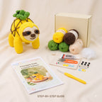Crochet kit for a pineapple sloth, featuring a finished yellow crochet sloth with green leaves, a cream box, yarn balls in yellow, green, brown, and beige, a crochet hook, a crochet pattern booklet, and other crafting accessories on a cream fabric background.