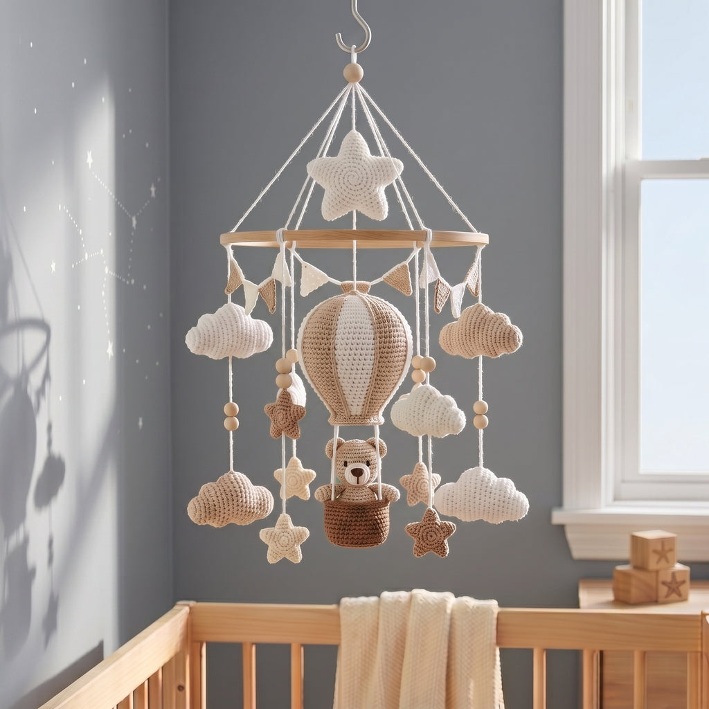 A crocheted baby mobile with a hot air balloon, teddy bear, stars, and clouds hanging in a wooden crib against a gray wall near a window.