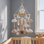 A crocheted baby mobile with a hot air balloon, teddy bear, stars, and clouds hanging in a wooden crib against a gray wall near a window.
