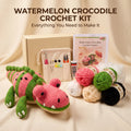 Watermelon Crocodile Crochet Kit displayed on a beige fabric background, featuring a crocheted watermelon crocodile toy, a beige gift box, a drawstring pouch with crochet hooks, a crochet pattern booklet, a tube of yarn adhesive, and multiple balls of yarn in pink, green, white, beige, and black.