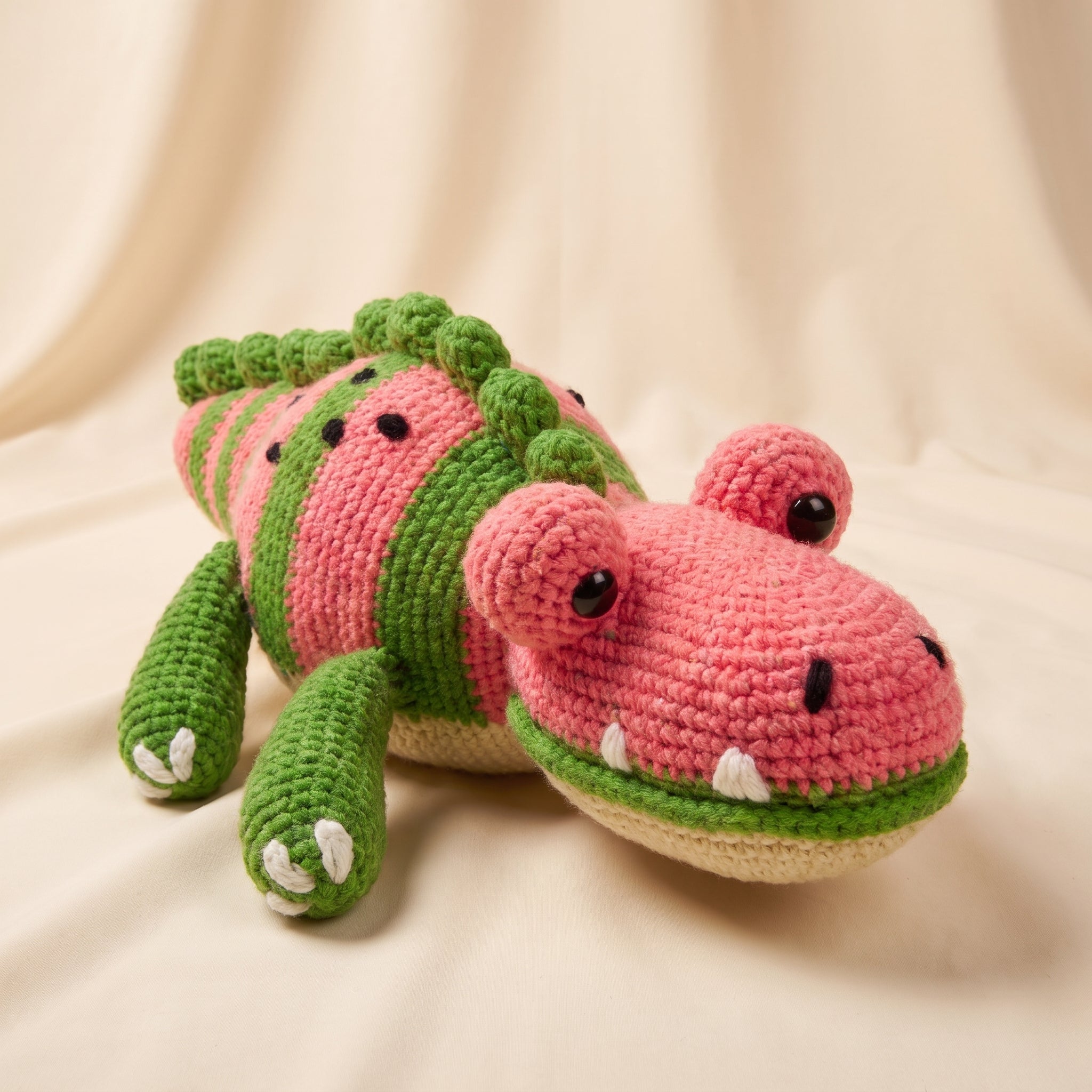 A crocheted plush toy shaped like a watermelon crocodile or alligator, with pink and green stripes, black bead eyes, white teeth, and small white claw details, photographed on a soft cream fabric background.