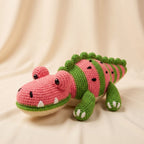 A crocheted plush alligator toy designed like a watermelon, featuring a pink and green striped body, black seed details, and a beige underbelly, set against a draped cream fabric background.