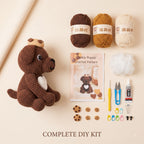 A crocheted dog DIY kit laid out on a warm beige background, including a brown crochet dog toy, three balls of yarn in brown, mustard, and cream, a crochet pattern booklet, white fiber, a yellow hook, blue and yellow scissors, a glue stick, small wooden cookie pieces, and colorful needle cases. Text at the bottom reads 'COMPLETE DIY KIT'.