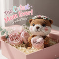 A crocheted plush toy bear with a floral crown and a pink heart reading “MOM,” nestled in a pink gift box with paper shred and pink flowers. Large text at the top reads “The Cutest Mom Ever! Crochet Hug!”.