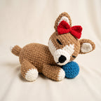 A crocheted plush puppy toy lying on a cream fabric background, holding a blue ball. The puppy is brown with white accents, a red bow on its head, and a black nose.