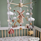 A crocheted baby mobile with a farm theme, hanging above a crib. The mobile features a wooden crossbar, white yarn clouds, a central windmill, a red barn, wheat stalks, stars, fence pieces, and hay bales in earthy brown, beige, and white tones.