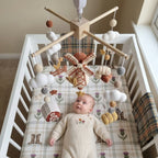A baby in a cream knit romper lies in a white crib, looking up at a wooden baby mobile with crocheted clouds, a windmill, and small autumn-themed hanging toys.