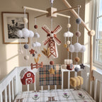 A rustic nursery crib setup featuring a handmade crocheted mobile with a windmill, clouds, stars, and a small red barn, hanging from a wooden arch above a white crib with matching plaid bedding.