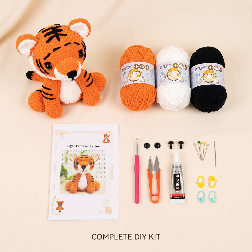 A DIY crochet kit laid out on a light beige background, including a crocheted tiger plush, three balls of yarn in orange, white, and black, a tiger crochet pattern booklet, a pink crochet hook, orange-handled scissors, a tube of glue, black buttons, yarn needles, and stitch markers. Text at the bottom reads 'COMPLETE DIY KIT'.