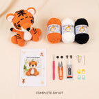 A DIY crochet kit laid out on a light beige background, including a crocheted tiger plush, three balls of yarn in orange, white, and black, a tiger crochet pattern booklet, a pink crochet hook, orange-handled scissors, a tube of glue, black buttons, yarn needles, and stitch markers. Text at the bottom reads 'COMPLETE DIY KIT'.