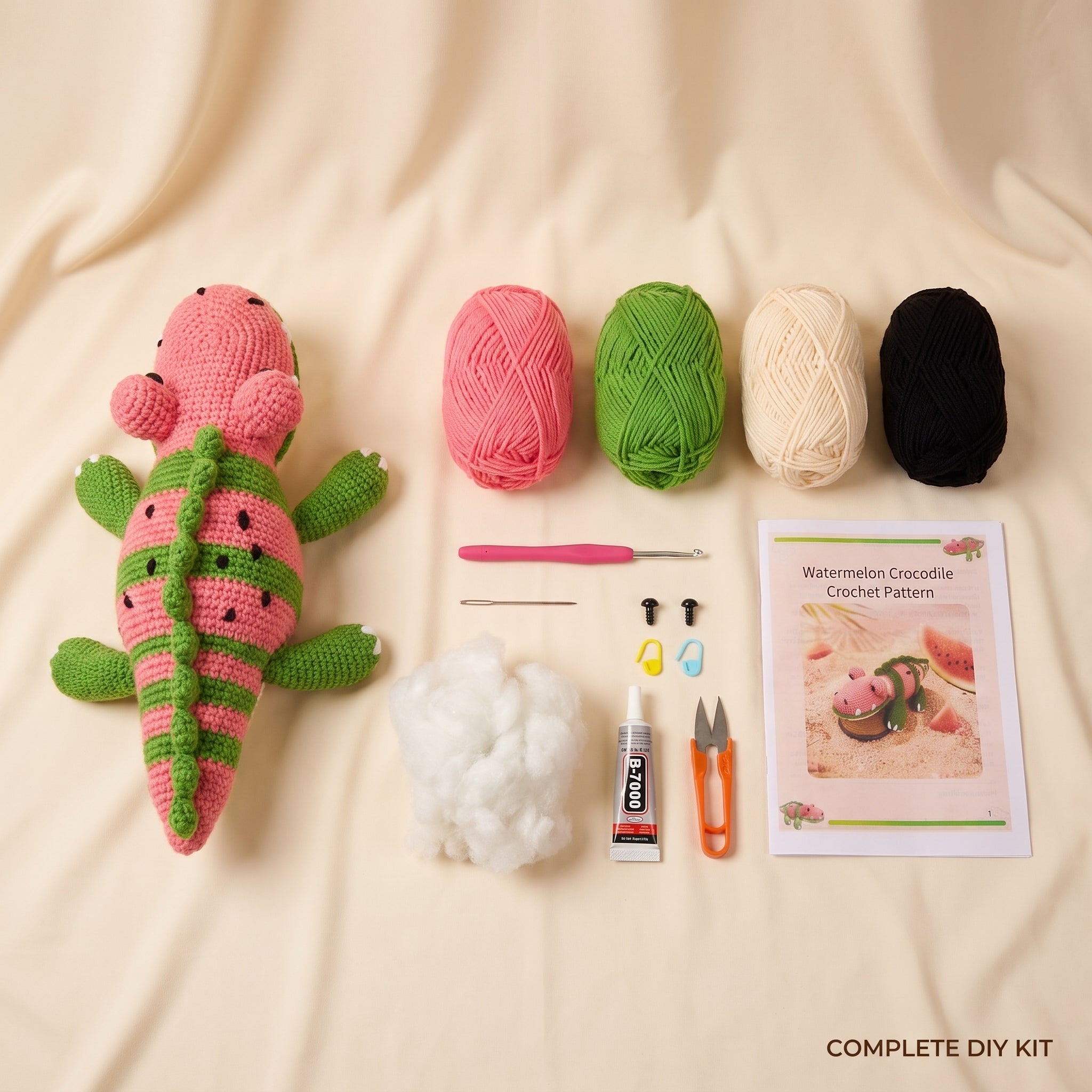 DIY crochet kit laid out on a cream fabric background, including a crocheted watermelon crocodile toy, four balls of yarn in pink, green, cream, and black, a crochet hook, needle, thread, glue, scissors, and a crochet pattern booklet.