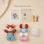 A flat-lay product presentation of a crochet kit titled 'EVERYTHING INCLUDED' on a warm beige background. The kit contains a finished crocheted fox toy in orange and blue, four balls of yarn in red, light blue, white, and pink, a ball of white stuffing, a beige drawstring pouch, a crochet hook, scissors, and a glue tube, alongside a printed instruction card.