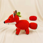 A crocheted plush fox toy in bright red, white, and brown, posed on a cream fabric backdrop with a small red ball of yarn behind it.