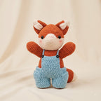 A crocheted plush toy fox wearing light blue overalls, sitting against a soft beige fabric backdrop.