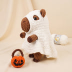 A crocheted plush toy dressed in a white ghost costume with brown ears, nose, and paws, standing on a beige fabric backdrop. A small orange pumpkin-shaped bag with a brown handle sits beside it, and a ball of white yarn is visible in the background.