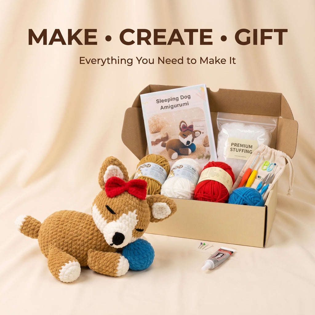 A craft kit set displayed on a beige fabric backdrop, featuring a crocheted sleeping dog toy with a red bow and blue ball, an open cardboard box containing yarn balls, a book titled 'Sleeping Dog Amigurumi,' a tube of paint, and other crafting supplies. Text at the top reads 'MAKE • CREATE • GIFT' and 'Everything You Need to Make It.
