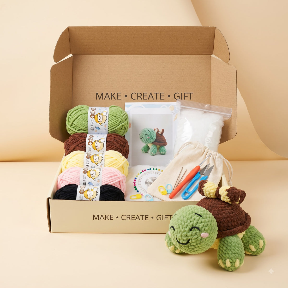 A crocheted turtle gift kit displayed in an open kraft cardboard box on a warm beige studio background. The kit includes multiple balls of yarn in green, brown, yellow, pink, and black, a small white pouch, a crochet hook set, and a finished green and brown crocheted turtle toy placed in front of the box.