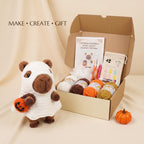 A crocheted plush bear dressed in a white ghost costume holding a small orange pumpkin is shown beside an open cardboard gift box containing a crochet pattern booklet, yarn balls, crochet tools, and a small pouch. The words 'MAKE • CREATE • GIFT' appear in the upper left on a beige draped backdrop.