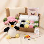 Crochet flower penguin plush toy kit displayed in a cardboard gift box with yarn balls, crochet hooks, scissors, and a pattern booklet, alongside a crocheted penguin plush wearing a pink flower hat on a beige fabric background.