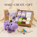 Crochet gift kit with a purple grape hippo plush toy, an open box of yarn balls, a crochet pattern booklet, poly fill, scissors, beads, and a drawstring bag on a beige fabric background. Text at the top reads 'MAKE • CREATE • GIFT'.