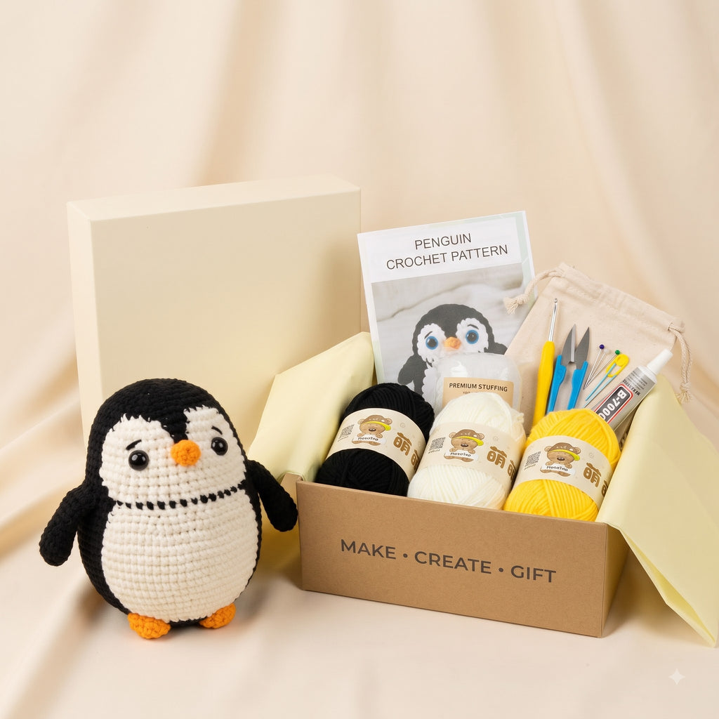 A crocheted penguin toy sits beside an open kraft gift box containing a crochet pattern booklet, black and cream yarn balls, yellow yarn, crochet hooks, needles, and a small pouch, on a beige fabric backdrop.