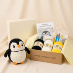 A crocheted penguin toy sits beside an open kraft gift box containing a crochet pattern booklet, black and cream yarn balls, yellow yarn, crochet hooks, needles, and a small pouch, on a beige fabric backdrop.