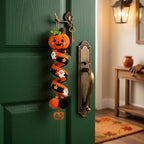 A crocheted Halloween keychain hanging from a brass doorknob on a dark green door. The keychain features a smiling orange pumpkin, a black and orange witch's hat with white ghosts, and a small jack-o'-lantern charm at the bottom, set against a cozy home entryway background.