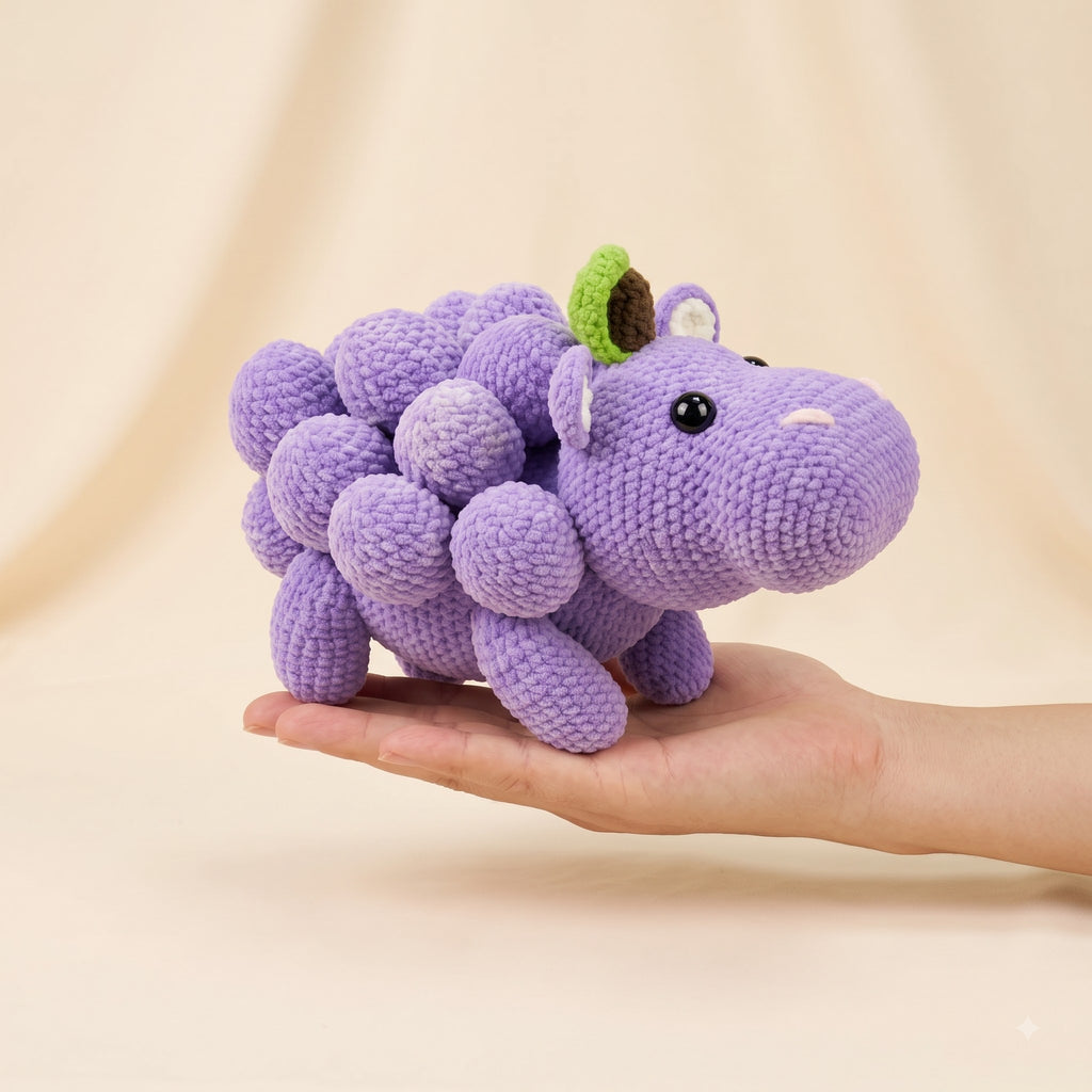 A small crocheted purple dinosaur toy with a green leaf on its head is held in a person's hand against a soft beige studio background.