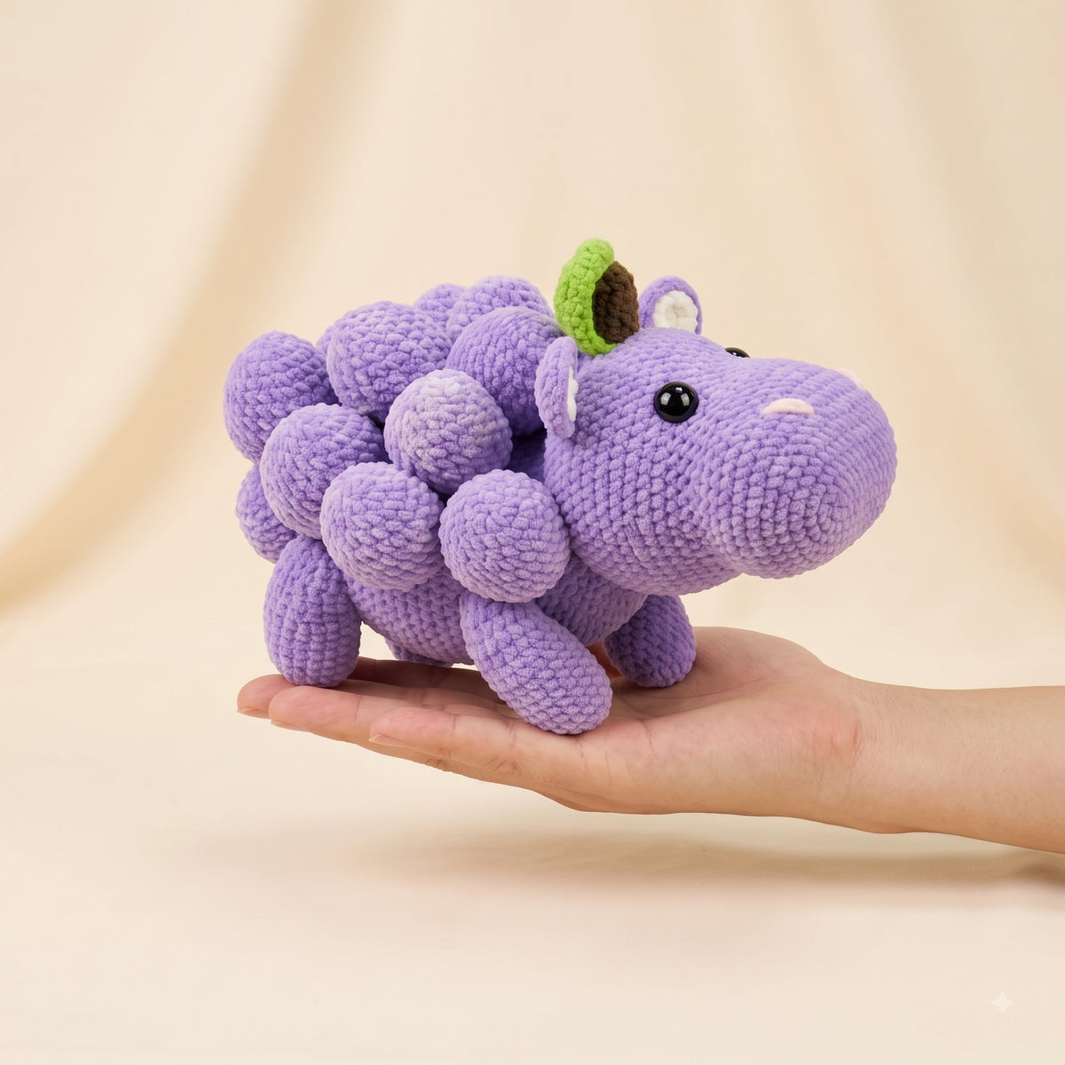A small crocheted purple dinosaur toy with a green leaf on its head is held in a person's hand against a soft beige studio background.