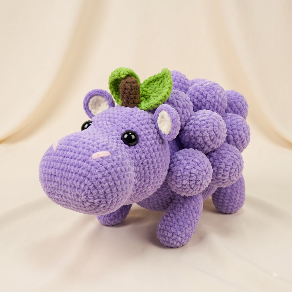 A crocheted plush toy shaped like a purple hippopotamus made of grape-like round shapes, with small green leaves and a brown stem on its head, photographed against a soft cream fabric backdrop.