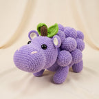 A crocheted plush toy shaped like a purple hippopotamus made of grape-like round shapes, with small green leaves and a brown stem on its head, photographed against a soft cream fabric backdrop.
