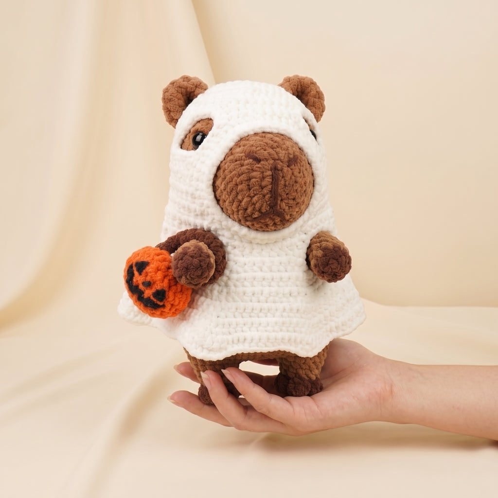 A crocheted capybara plush toy wearing a white ghost costume and holding an orange pumpkin, being held in a person's hand against a soft beige fabric backdrop.