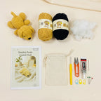 Crochet puppy kit laid out on a light beige background, including a finished yellow crochet puppy plush, a black yarn ball, white stuffing, a printed instruction booklet, a cream drawstring bag, a yellow crochet hook, orange-handled scissors, a tube of PTFE glue, and small sewing needles and stitch markers.