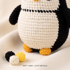 A close-up of a crocheted penguin toy with a cream body, black wings and back, and yellow feet and beak. Two small yarn balls, one matching the toy's cream body and one black, and one yellow yarn ball sit on a soft cream fabric surface.
