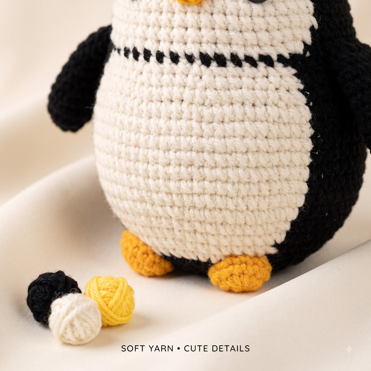 A close-up of a crocheted penguin toy with a cream body, black wings and back, and yellow feet and beak. Two small yarn balls, one matching the toy's cream body and one black, and one yellow yarn ball sit on a soft cream fabric surface.