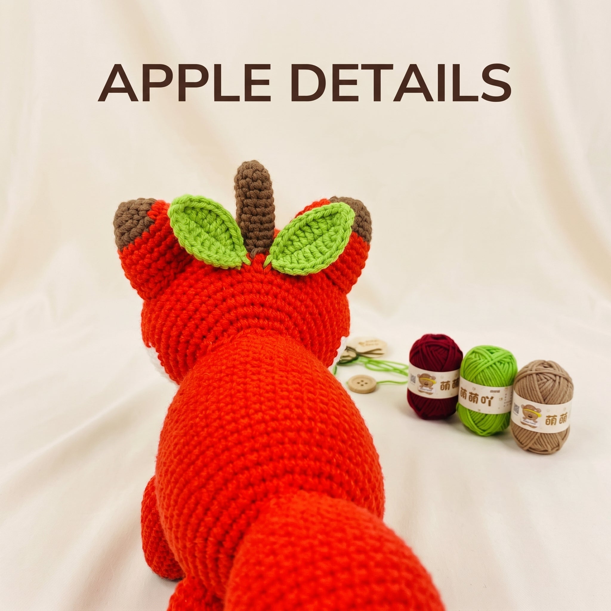 A crocheted apple-shaped fox or fox-apple toy shown from the back on a cream fabric background, with the text 'APPLE DETAILS' above it and three yarn balls and buttons to the right.